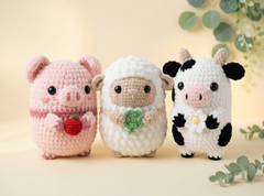 Three crocheted animal plush toys are lined up against a soft beige background with decorative green leaves. The left toy is a pink pig holding a red apple, the middle is a white sheep with a green clover, and the right is a black-and-white cow with a white flower.