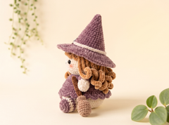 A small crocheted doll with curly brown hair, wearing a purple witch hat and matching dress, posed on a warm cream background with soft green plant leaves in the corners.