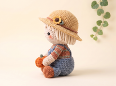 Crocheted amigurumi doll of a girl farmer wearing a straw hat with a sunflower decoration, holding a small orange pumpkin, set against a warm beige background with a eucalyptus leaf branch.