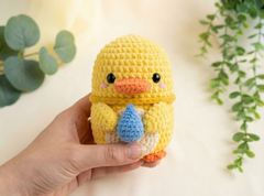 A hand holding a small crocheted yellow duck toy with an orange beak, black eyes, and a blue teardrop accessory, set against a soft cream background with green plant leaves.