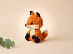 A crocheted fox toy sits centered on a warm beige background, with a small green leaf sprig in the lower left corner. The fox is orange with white and black details.