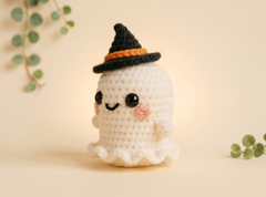 A cute crocheted ghost doll wearing a black witch hat with an orange band, set against a warm beige background with soft eucalyptus leaves.