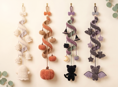 Four knitted Halloween hanging ornaments displayed against a warm cream background with eucalyptus leaves in the corners. The ornaments include a ghost, pumpkin, witch hat and cat, and bat designs in neutral cream, orange, purple, and gray tones.