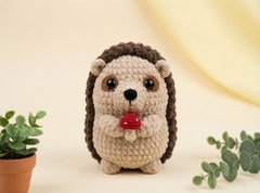 A crocheted hedgehog toy holding a small red mushroom, centered against a soft cream background with decorative greenery at the sides.