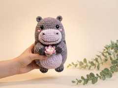 A crocheted hippo plush toy being held by a hand, featuring a dark gray body, light purple snout and belly, pink cheeks, and a small pink flower on the front, with soft-focus green leaves in the background.