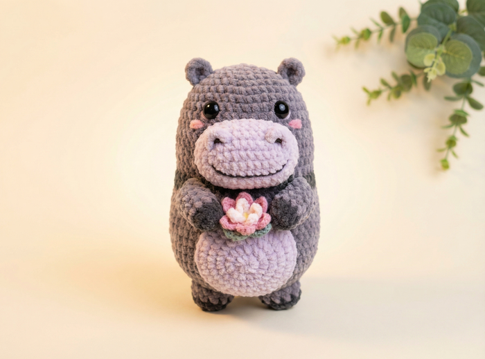 A crocheted hippo plush toy in dark gray and light lavender, holding a small pink flower, set against a warm cream background with a decorative green plant in the upper right corner.