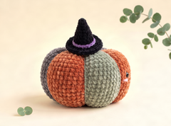 A crocheted pumpkin-shaped amigurumi doll with orange, gray, and sage green panels, wearing a black witch hat with a purple band, photographed on a warm cream background with eucalyptus leaves.