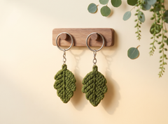Two olive green crocheted leaf keychains hanging from silver metal rings on a small wooden display stand, with soft eucalyptus leaves in the background.