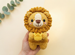 A hand holding a crocheted lion plush toy with a golden-yellow mane, cream-colored face, and a small yellow crown, set against a warm beige background with a decorative eucalyptus sprig in the corner.