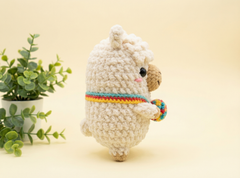 A crocheted llama plush toy in cream white yarn, featuring a colorful rainbow-striped collar and holding a matching multicolor ball, set against a warm beige background with a small green potted plant on the left.