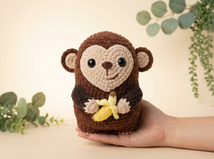 A crocheted plush monkey toy in brown and beige, holding a small yellow banana, displayed on a person's hand against a warm beige background with soft green plant leaves.