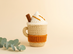 Beige crochet coffee cup topped with cream, caramel drizzle chains and a brown cinnamon stick.