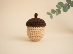 A crocheted acorn ornament with a dark brown cap and stem, and a beige textured body, displayed on a warm beige background with a soft-focus eucalyptus branch in the upper right.