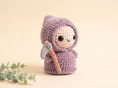 A small crocheted amigurumi doll wearing a lavender-purple hooded outfit, holding a gray and peach pickaxe, set against a warm beige background with a sprig of green leaves in the lower left corner.