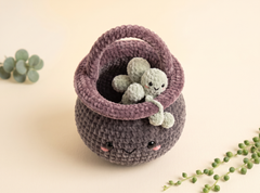 A handmade crocheted basket in dark gray and dusty purple, featuring a small plush octopus toy inside, styled with green plant stems on a warm cream background.