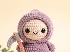 A cute crocheted doll wearing a lavender-purple hooded costume and holding a small gray scythe, set against a soft beige background.