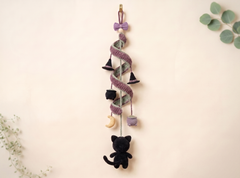 A hanging Halloween-themed yarn decoration featuring a black plush cat, small witch hats, a hanging cauldron, a crescent moon, and purple and gray wrapped yarn, displayed against a warm cream background with subtle green leaves in the corners.
