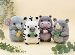 Four crocheted animal plush toys arranged together on a light beige background with decorative green leaves. The toys include a gray elephant holding a yellow banana, a white zebra with black stripes holding a green leaf, a lavender hippo holding a pink lotus, and a gray rhinoceros holding a green leaf and cream horn.