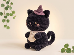 A crocheted black cat plush toy wearing a purple witch hat and a cream bib, posed against a warm beige background with soft green plant leaves in the corners.