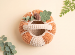 A crocheted pumpkin-shaped pet bed with a white handle and green leaf accents, photographed on a warm cream background with decorative eucalyptus leaves in the corners.