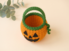 A crocheted pumpkin-shaped candy bucket with a green handle and stem, featuring a black embroidered jack-o'-lantern face, set against a warm beige background with a decorative eucalyptus sprig in the corner.