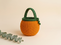 A crocheted mini pumpkin bag with a green handle and stem detail, styled on a warm beige background with a sprig of eucalyptus in the foreground.