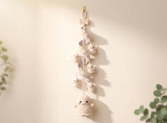 A cream-colored crocheted ghost hanging ornament with a spiral ribbon detail, small smiling ghost charms, and a bow at the top, hanging on a warm beige wall with soft shadows.