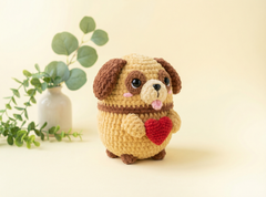 A crocheted plush dog toy with brown ears and paws, a cream-colored body, and a red heart on its front paws, standing against a warm cream background with a small white vase and green eucalyptus leaves on the left.