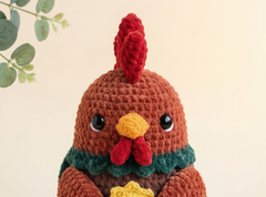 A crocheted toy chicken with a brown body, red comb and wattle, yellow beak, and dark green neck trim, photographed against a soft cream background with a small leafy branch in the upper left.