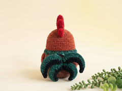 A crocheted rooster toy shown from the back, featuring a terracotta brown body, a bright red comb and wattle, and dark green and black tail feathers, set against a warm cream background with a small green plant in the corner.