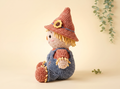 A crocheted scarecrow doll sitting on a warm beige background, wearing a brown hat with a yellow flower, a blue-gray overalls outfit, and yellow hair.
