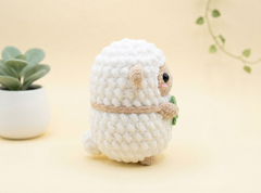 A small white crocheted plush sheep toy with beige ears, a beige band around its neck, and a tiny green leaf in its front paw, standing on a soft cream background with a potted plant on the left.