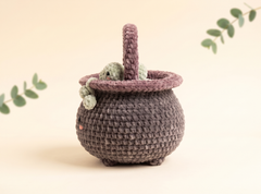 A crocheted cauldron-shaped basket in dark gray with a dusty purple handle and rim, featuring small green yarn creatures peeking out from the side, set against a warm beige background with decorative eucalyptus leaves.