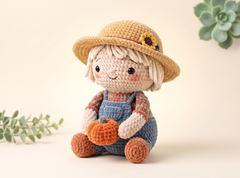 A crocheted doll styled as a harvest girl, wearing a straw hat with a sunflower, blue overalls, and holding a small orange pumpkin, set against a warm beige background with soft green plant accents.