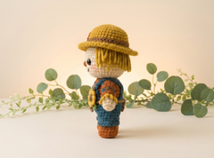 A crocheted scarecrow doll stands on a light beige surface with eucalyptus branches in the background. The doll wears a yellow hat with a brown band, a cream face and hair, a blue top, and brown pants, and holds a small sunflower.