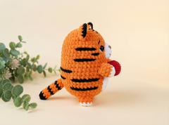 A crocheted orange tiger toy with black stripes, white paws and face, holding a red apple, set against a warm beige background with green eucalyptus leaves on the left.