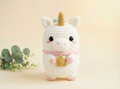 A cute crocheted unicorn plush toy in white with a gold horn, pink collar, and pink hooves, standing against a soft beige background with a small green plant on the left.