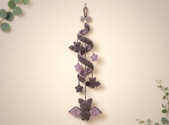 A handmade knitted bat-shaped hanging decoration with purple wings and a purple bow at the top, shown against a warm cream background with subtle green leaves in the corners.