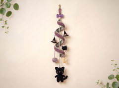 A hanging Halloween ornament made from a twisted purple and gray ribbon, decorated with small witch hats, a cauldron, a moon, and a black cat plush at the bottom, shown against a warm beige wall with green leaves in the corners.