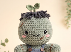 A close-up of a crocheted plush character with a round pale green head, dark gray yarn hair, pink cheeks, and blue overalls against a soft beige background.
