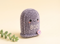 A cute, hand-crocheted ghost toy in lavender purple, featuring embroidered eyes, a smiling mouth, the word 'BOO' in white, and a small pink flower on the side. It is styled on a warm beige background with a small green succulent sprig in the foreground.