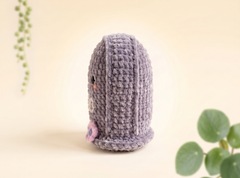 A small crocheted plush toy shaped like a cute character, shown from the back on a warm cream background with soft green plant leaves in the corners.