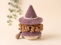 A crocheted gnome doll with a purple pointed hat, brown curly hair, and a cream-colored base, styled on a warm beige background with a hanging eucalyptus branch.