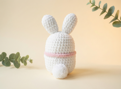 A white crocheted bunny plush toy with long ears and a pink band around its neck, shown from the back on a warm beige background with decorative eucalyptus branches.