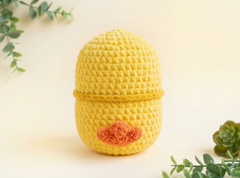 A yellow crocheted egg-shaped container with a small orange textured accent on the front, styled with green plants on a light cream background.