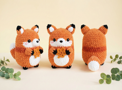 Three crocheted fox toys are displayed on a light beige surface against a cream background, showing front, back, and side views. The foxes are orange with white and black details, and small green leaves are scattered around them.
