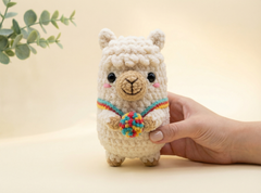 A small crocheted alpaca plush toy with a colorful rainbow scarf and multicolored pom-pom accessory, held by a human hand against a soft beige background.