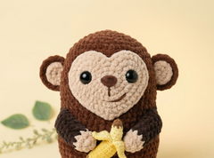 A crocheted plush monkey toy holding a yellow banana, centered against a soft beige background with a small green leafy sprig at the lower left.