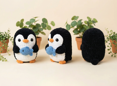 Three crocheted penguin toys arranged on a light beige surface with small potted green plants in the background. Two penguins face forward and hold a blue fish, while one penguin is shown from the back.