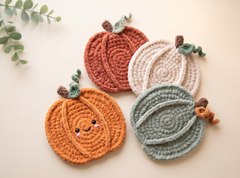 Four crocheted pumpkin-shaped coasters arranged on a light beige background, with a small green leafy sprig in the top left corner. The coasters are orange, rust red, cream, and sage green, with one featuring a cute embroidered face.